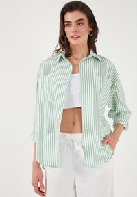 Green and white vertically striped button-up shirt with a relaxed fit, two chest pockets, and rolled sleeves, worn over a white crop top.
