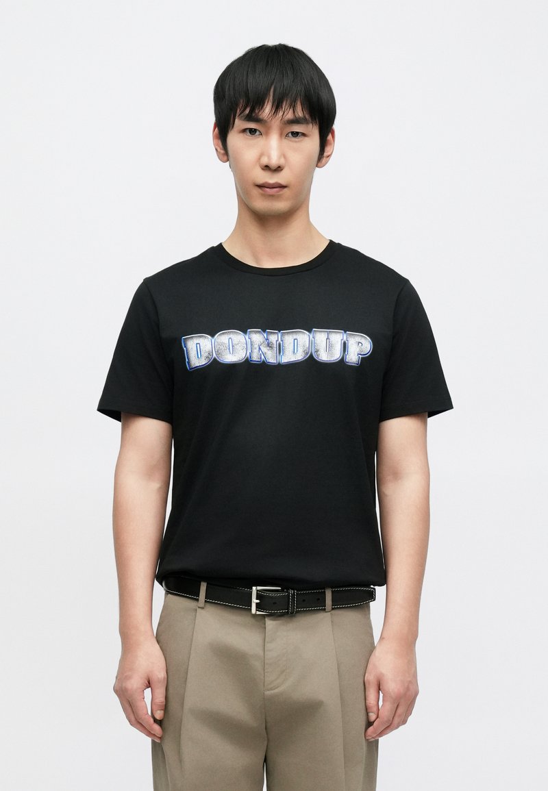 Μαύρο βαμβακερό μπλουζάκι με τη λέξη "DON DUP"  σε έντονα, μεταλλικό μπλε γράμματα. Φορεμένο με μπεζ παντελόνια και μαύρη ζώνη με λευκή ραφή.