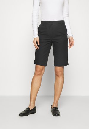 Frau trägt schwarze, knielange, maßgeschneiderte Shorts, weißes Langarmshirt und schwarze Loafers, steht vor weißem Hintergrund.