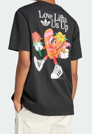 T-shirt en coton noir avec des graphiques de dessins animés colorés au dos, accompagnés d'un slogan, d'accents roses et d'un motif de baskets blanches.