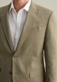 Hellbrauner Leinenblazer mit Reverskragen, einem einzelnen Knopf und einer Brusttasche, über einem weißen Leinenhemd mit sichtbaren Knöpfen getragen.