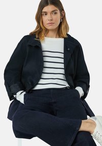 Marineblauwe waterdichte jas met een relaxte pasvorm, gedragen over een wit en marineblauw gestreept trui. Donker denim broeken maken de look compleet.