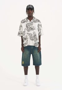 Korte mouw witte shirt met een zwart drakenprint, gecombineerd met denim shorts, witte sneakers en een zwarte pet met zilveren accenten.