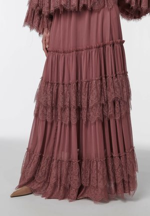 Personne vêtue d'une longue robe mauve à volants en dentelle superposés, debout avec une main le long du corps et des chaussures à bouts pointus visibles.