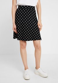 Mujer vestida con una falda negra con un patrón de diamantes blancos, una blusa blanca y zapatillas de plataforma blancas, de pie contra un fondo liso.