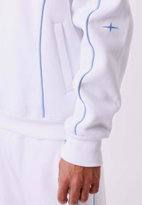 Witte sweatshirt met lichtblauwe piping langs de naden. Bevat ribgebreide manchetten en een zijzak. Gladde textuur met een minimalistisch ontwerp.