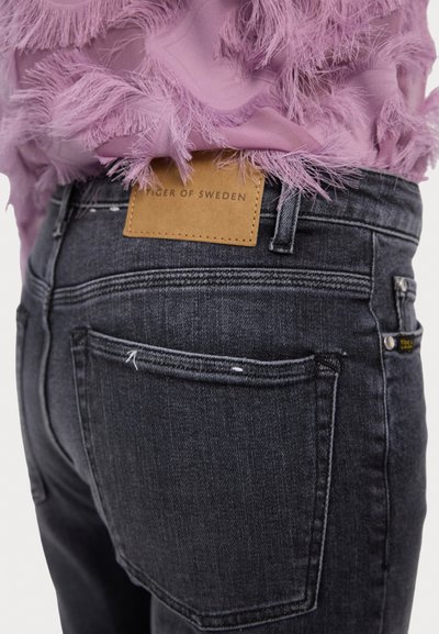 Jeans de mezclilla gris oscuro con una etiqueta de cuero que dice "Tiger of Sweden", con detalles de costura y una ligera textura desgastada.