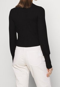 Schwarzes, geripptes Langarmtop, kombiniert mit hellbeigen, hochtaillierten Hosen, die eine strukturierte Oberfläche und dezente Rückendetails aufweisen.