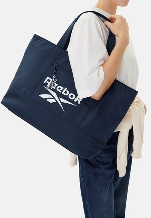 Osoba nosząca białą koszulę i dżinsy, niosąca dużą granatową torbę Reebok na ramię, z przednią kieszenią na suwak.