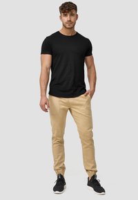 Sort tætsiddende t-shirt, beige joggingbukser med elastiske manchetter og sidelommer. Båret med sorte træningssko. Neutral baggrund.