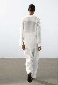 Camicia bianca trasparente con maniche lunghe e spacchi laterali, abbinata a pantaloni bianchi larghi. Tessuto morbido, design minimalista e vestibilità rilassata.