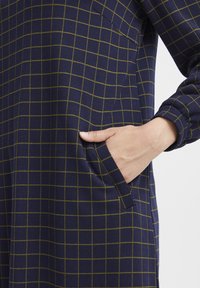 Marineblaues Kleid mit einem Karomuster in Gelbgrün, mit einer Seitentasche und langen Ärmeln, aus einem weichen Stoff gefertigt.