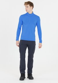 Langærmet, lynlås skjorte i lys blå, parret med mørke cargo bukser. Stoffet ser strækbart ud; outfittet er designet til aktiv brug.