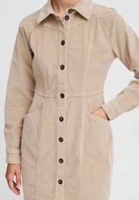 Beige kordrocksklänning med spetsig krage, knäppning framtill, två fickor och svarta knappar, i ett texturerat, ribbat tyg.