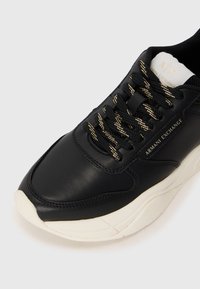 Baskets en cuir noir avec des accents dorés sur les lacets, dotées d'une semelle en caoutchouc blanche et d'un col de cheville rembourré. Logo Armani Exchange visible.