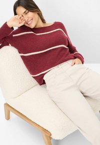 Burgunderfarbener Strickpullover mit cremefarbenen Streifen, kombiniert mit beigen Chinos. Sitzt auf einem weißen, flauschig strukturierten Stuhl mit Holzbeinen.