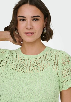 Femme aux cheveux bruns courts portant des boucles d'oreilles argentées en créoles et un haut en tricot vert menthe avec des motifs complexes, posant avec la main derrière la tête.