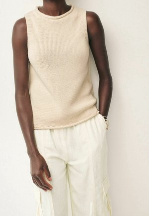 Tummaihoinen henkilö, jolla on päällään hihaton beige neulepaita ja väljät valkoiset housut, joissa on taskut, seisoo yksinkertaisen vaalean taustan edessä.