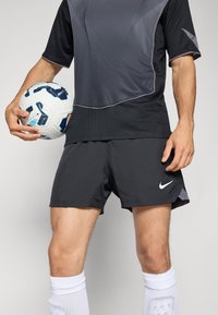 Mannelijke voetballer in zwarte sportkleding met een wit-blauwe voetbal aan zijn zijde, gekleed in witte sokken en korte broek met een wit swoosh-logo.