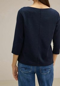 Námořnická modrá, strukturovaná blůza s tříčtvrtečními rukávy, která má čtvercový výstřih a šev na zadní straně, doplněná světlými modrými džínami.