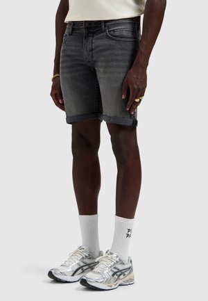 Man draagt donkergrijze opgerolde denimshorts, witte sokken met tekst, zilveren hardloopschoenen en een wit overhemd, staand tegen een effen achtergrond.