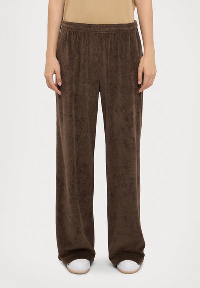 Bruine, wijde fleece broeken met een elastische tailleband. Zachte textuur en vloeiende stijl, gecombineerd met witte sneakers onderaan.