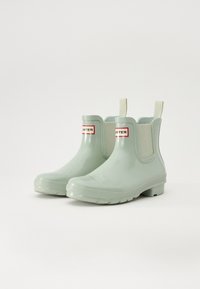 Bottines en caoutchouc vertes avec des panneaux latéraux élastiques, des semelles texturées et un patch logo rouge et noir à l'avant. Fini lisse, bout arrondi.