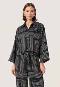 Cămașă neagră scurtă cu un model geometric alb, cu mâneci lungi, guler și nasturi în față. Asortată cu pantaloni cu model similar.