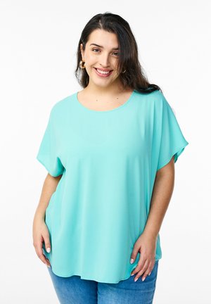Lys turkise kortærmede top i et blødt, draperende materiale med rund halsudskæring. Matchet med blå denim jeans.