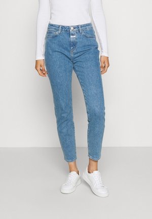 Personne portant un jean taille haute bleu clair, un haut blanc à manches longues et des baskets blanches, debout sur un fond uni.