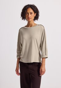 Eine Frau steht da und trägt ein beige-farbenes Langarmoberteil mit schwarzen Streifen an den Ärmeln und dunkelbraune Hosen vor einem weißen Hintergrund.