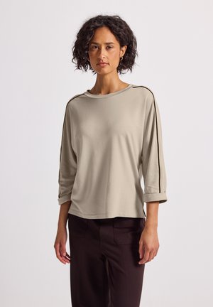 Femme debout portant un haut beige à manches longues avec des rayures noires sur les manches et un pantalon marron foncé, le tout sur fond blanc.