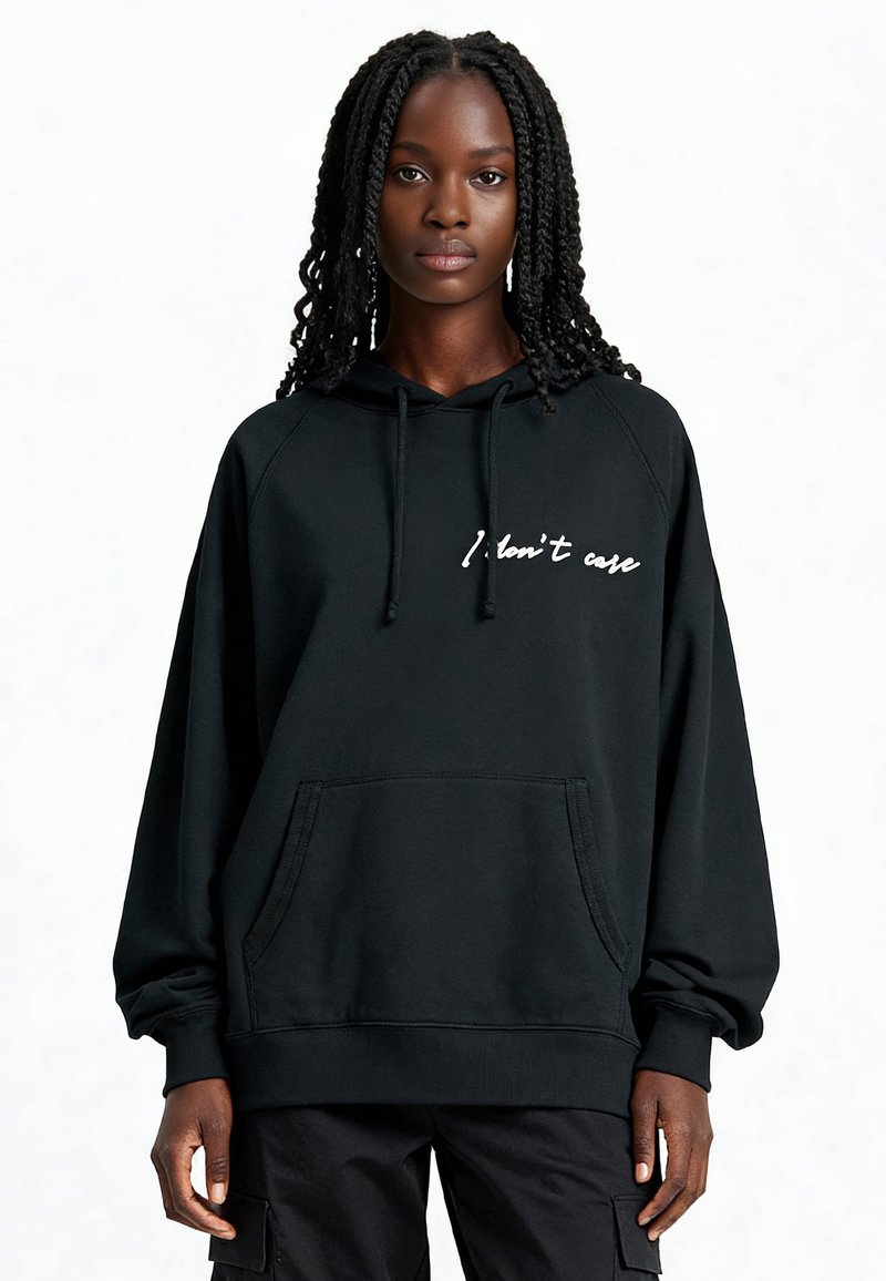 Sweatshirt à capuche noir avec poche avant, comportant un texte blanc en cursive "I don't care" sur la poitrine. Tissu doux, coupe décontractée, et poignets côtelés.