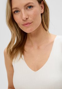 Weißes, geripptes Tanktop mit V-Ausschnitt, das eine figurbetonte Form und eine glatte Textur aufweist, vor einem neutralen Hintergrund.