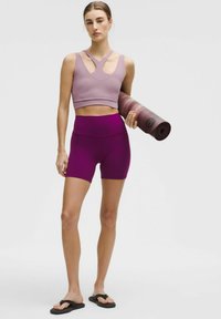 Lila sportbh med skärdesign, kombinerad med högmidjade lila shorts; håller i en rullad yogamatta med gradientdesign. Svarta flip-flops.