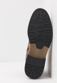 Zapato de cuero marrón con una suela de goma texturizada. La suela presenta patrones en zigzag y en bloques para mejorar la tracción. No hay logotipos reconocibles.