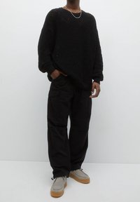 Zwarte gebreide trui met een gestructureerd weefpatroon, gecombineerd met losse zwarte cargobroeken en lichtgrijze schoenen met een gumzool.