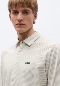 Ilgomis rankovėmis, kreminės ir baltos spalvos dryžuota marškiniai su klasikiniu apykakle, kairiojo krūtinės pusės juodo „BOSS“ logotipo ir lygaus audinio tekstūros.