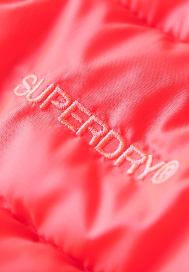 Der leuchtend rosa Jackenstoff verfügt über ein gesticktes Logo aus Pfirsichfaden mit der Aufschrift "SUPERDRY". Glatte Textur mit leicht glänzendem Finish.