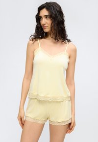 Femme aux cheveux bouclés foncés portant un caraco jaune pâle et un short assorti à bordure en dentelle, debout devant un fond clair uni.