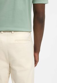 Mand iført lysegrøn skjorte stoppet ned i cremefarvede bukser, vist fra taljen til midt på låret med en arm synlig ved siden.