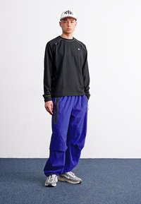 Camisa negra de manga larga, pantalones cargo azules de corte holgado, zapatillas grises y una gorra blanca con texto. Tejido suave y características de diseño prácticas.