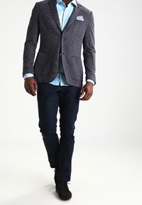 Grijze herringbone blazer met twee zakken, gecombineerd met een lichtblauw shirt en marineblauwe broek; zwarte schoenen maken de outfit compleet.