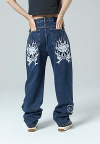 Jeans de pierna ancha de mezclilla en azul oscuro que presentan grandes gráficos de calaveras con llamas a cada lado, y un detalle de logo circular en el dobladillo.