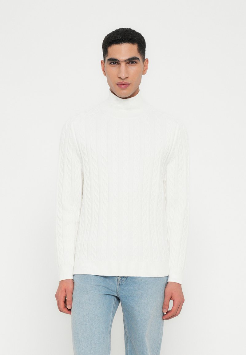 Jersey de cuello alto de punto blanco que presenta un diseño texturizado, con mangas largas y una silueta ajustada, combinado con unos vaqueros azul claro.