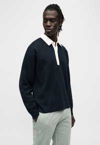 Μπλε σκούρο πουλόβερ με λευκό γιακά, διαθέτει σειρά κουμπιών και φαρδιά μανίκια. Φοριέται με ανοιχτό πράσινο παντελόνι από κόρντουρα. Λεία υφή.