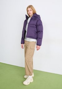 Vijolična napihljena jakna s kapuco, zadrgo in elastičnimi manšetami, v kombinaciji s bež cargo hlačami in kremnimi supergami na zeleni podlagi.