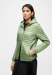 Γυναίκα που φορά ανοιχτό πράσινο μπουφάν με κουκούλα, μαύρο τοπ και μαύρη πλισέ φούστα, με το ένα χέρι στην τσέπη του μπουφάν, ουδέτερο φόντο.