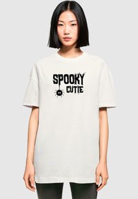 Krēmveida oversize T-krekls, kas izgatavots no kokvilnas, ar melnu grafiku, kas lasa "SPOOKY CUTIE" ar zirnekli, īsām piedurknēm un apaļu kakla izgriezumu.
