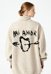 Giacca oversize in pile beige con collo alto e grafica nera "Mi Amor" con un disegno di cuore e freccia sul retro.